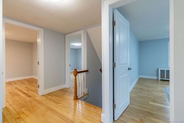 corridor with light wood-type flooring, a textured ceiling, baseboards, and an upstairs landing