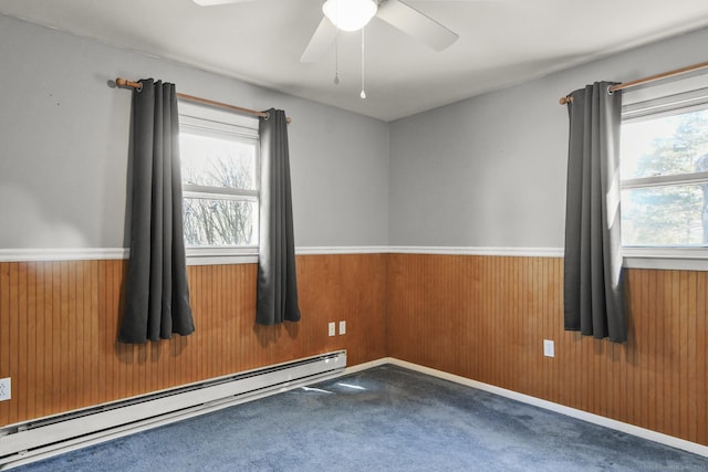 carpeted empty room with a baseboard radiator, wooden walls, ceiling fan, and wainscoting