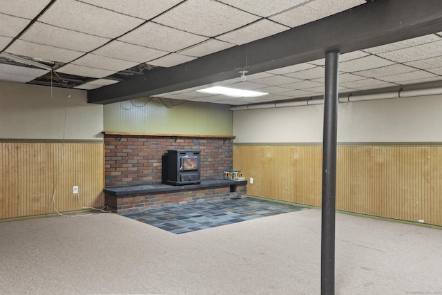 below grade area with a drop ceiling, wooden walls, carpet flooring, wainscoting, and a wood stove