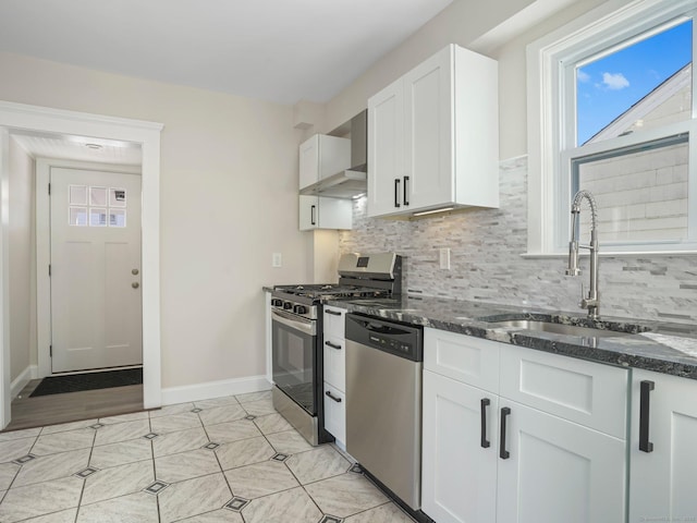 kitchen featuring dark stone counters, white cabinets, stainless steel appliances, wall chimney exhaust hood, and a sink