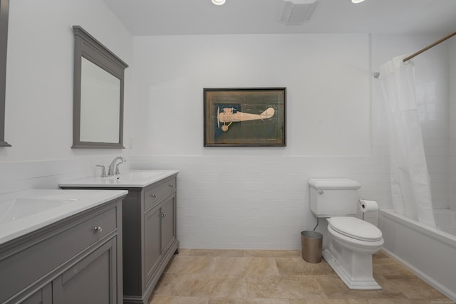 bathroom featuring toilet, two vanities, wainscoting, shower / bath combination with curtain, and a sink