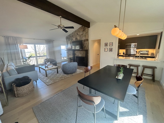 dining area with a stone fireplace, light wood-style flooring, vaulted ceiling with beams, and ceiling fan