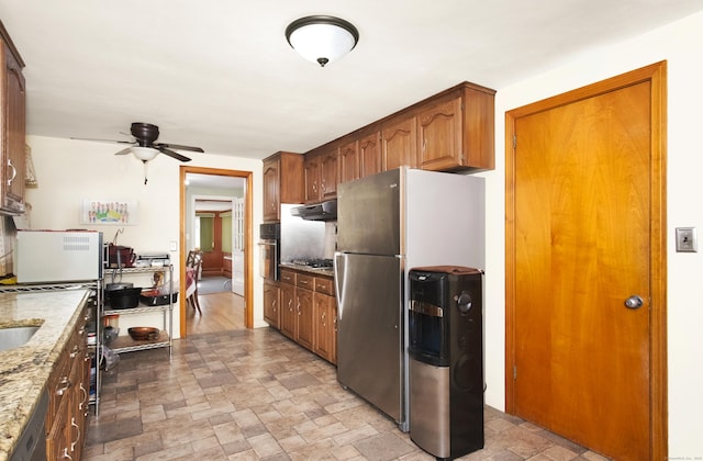 kitchen with stone finish floor, stainless steel appliances, brown cabinetry, light stone countertops, and ceiling fan