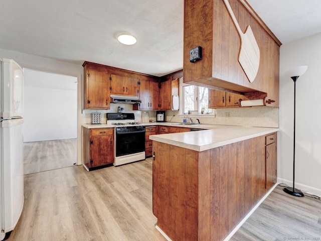 kitchen with brown cabinetry, freestanding refrigerator, light countertops, gas range oven, and under cabinet range hood