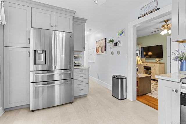 kitchen with gray cabinetry, light wood-type flooring, light stone counters, stainless steel fridge, and a ceiling fan