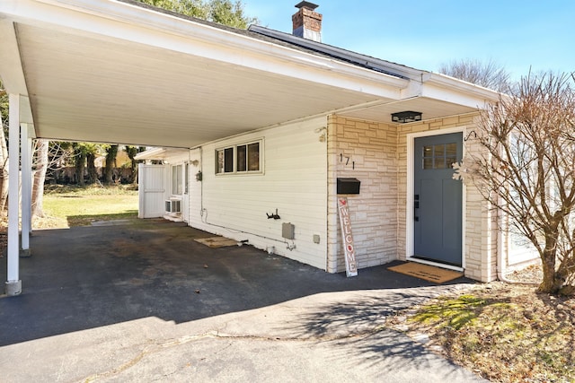 entrance to property featuring driveway, stone siding, an attached carport, and a chimney