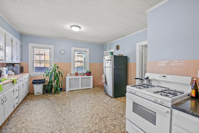 kitchen with white gas range oven, white cabinetry, freestanding refrigerator, crown molding, and wainscoting