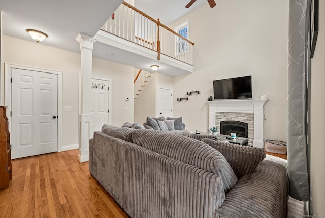 living room with ceiling fan, light wood-style floors, a stone fireplace, a towering ceiling, and stairs