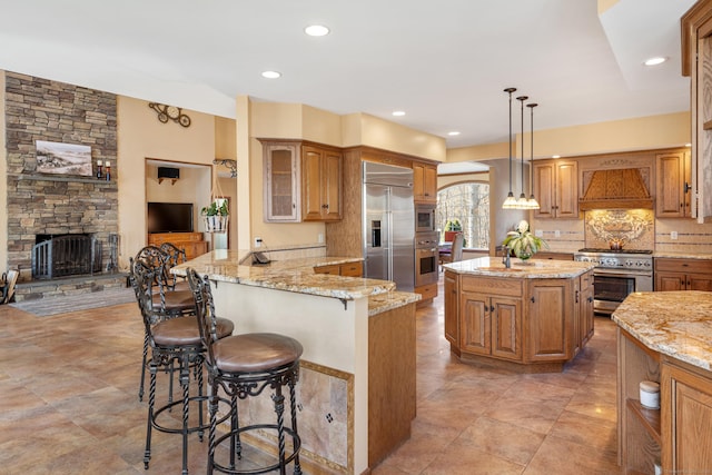 kitchen with backsplash, light stone countertops, premium range hood, built in appliances, and a breakfast bar