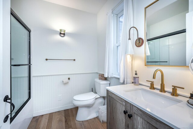 bathroom with wood finished floors, wainscoting, toilet, vanity, and bath / shower combo with glass door