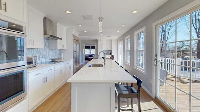 kitchen featuring a sink, backsplash, white cabinetry, stainless steel appliances, and wall chimney range hood