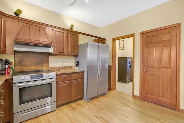 kitchen featuring range hood, light stone counters, brown cabinetry, light wood-style flooring, and appliances with stainless steel finishes