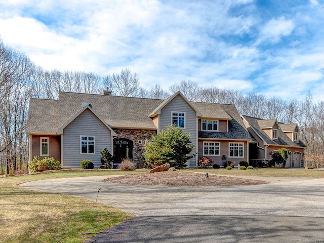 view of front facade featuring a front lawn, driveway, stone siding, roof with shingles, and a chimney