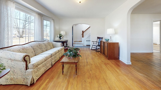 living area featuring arched walkways, stairway, light wood-style floors, and a baseboard radiator