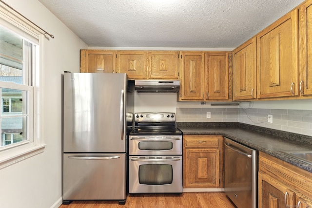 kitchen with light wood finished floors, dark countertops, under cabinet range hood, brown cabinetry, and stainless steel appliances