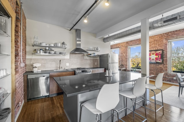 kitchen with dark countertops, brick wall, wall chimney range hood, appliances with stainless steel finishes, and a sink