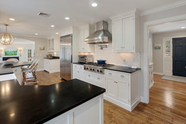 kitchen featuring light wood-style floors, crown molding, appliances with stainless steel finishes, dark countertops, and wall chimney range hood