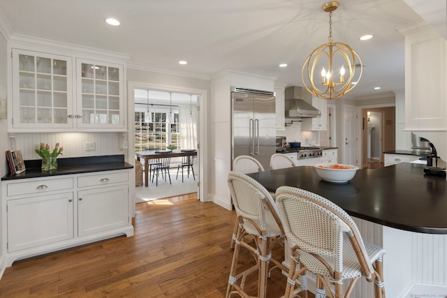 kitchen featuring ornamental molding, stainless steel appliances, white cabinetry, dark countertops, and wall chimney range hood