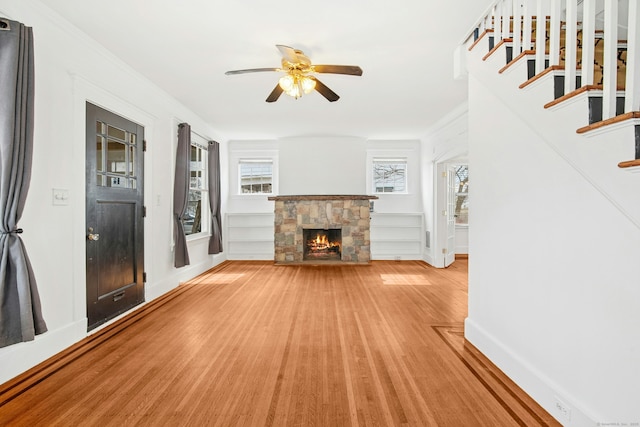 unfurnished living room with stairway, a ceiling fan, baseboards, light wood-style flooring, and a stone fireplace