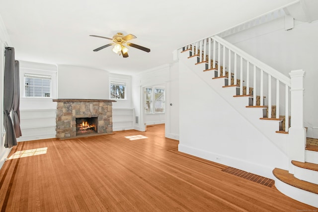 unfurnished living room featuring baseboards, light wood-type flooring, stairs, a stone fireplace, and a ceiling fan