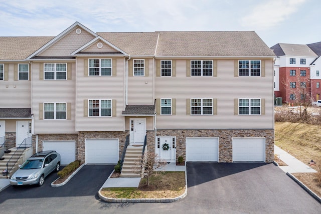 multi unit property featuring stone siding, driveway, a garage, and roof with shingles