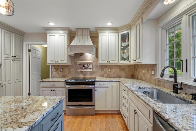 kitchen featuring light wood finished floors, light stone countertops, custom exhaust hood, stainless steel appliances, and a sink