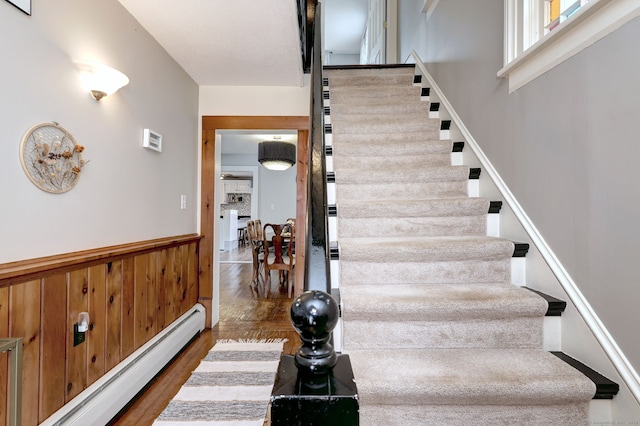 staircase with wood finished floors, wood walls, baseboard heating, and wainscoting