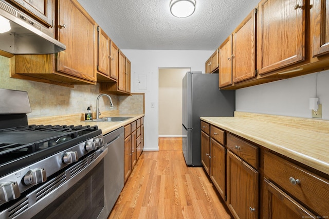 kitchen with a sink, light countertops, appliances with stainless steel finishes, under cabinet range hood, and brown cabinets