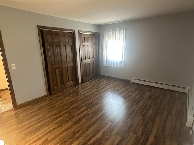 unfurnished bedroom with dark wood finished floors, baseboards, a baseboard heating unit, and multiple closets