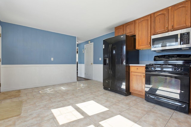 kitchen featuring light countertops, light tile patterned floors, wainscoting, brown cabinets, and black appliances