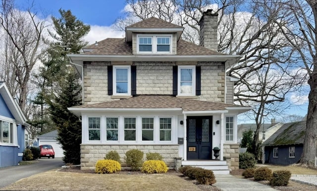 american foursquare style home featuring stone siding, french doors, roof with shingles, an outdoor structure, and a chimney