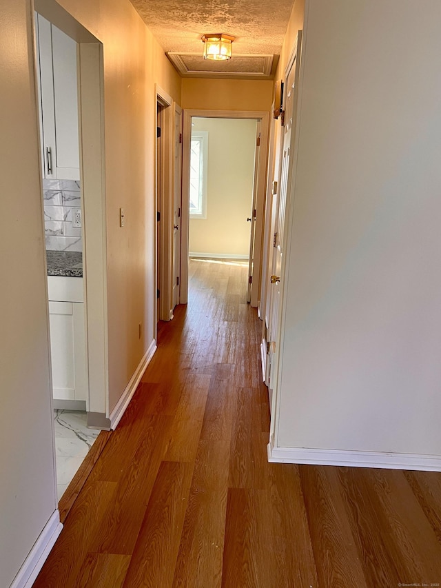 hall featuring attic access, light wood-style flooring, baseboards, and a textured ceiling