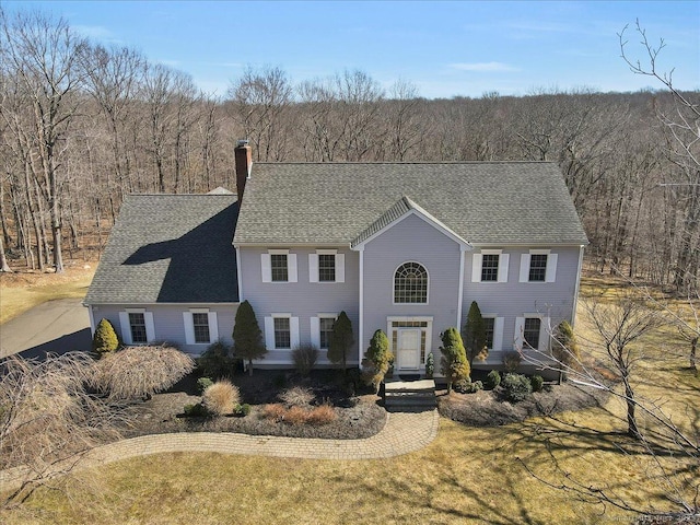 colonial inspired home featuring a chimney, a forest view, and a shingled roof