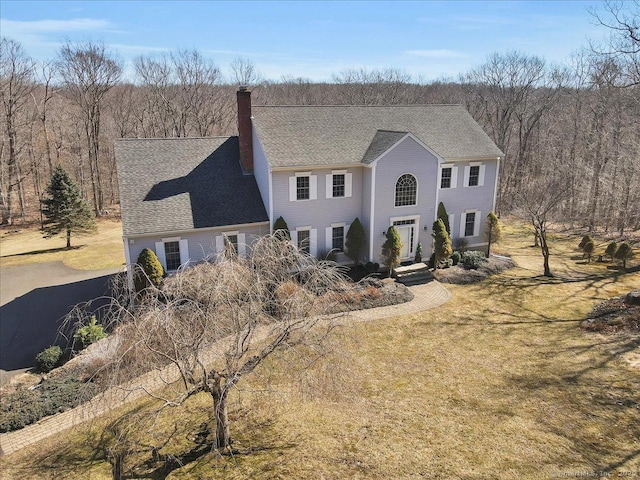 view of front of house featuring a shingled roof, a front lawn, a wooded view, and a chimney