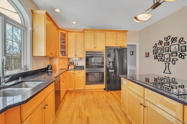 kitchen with dark stone countertops, a sink, black appliances, glass insert cabinets, and light wood-type flooring