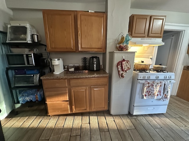 kitchen with under cabinet range hood, stainless steel microwave, dark countertops, white gas stove, and light wood-style floors