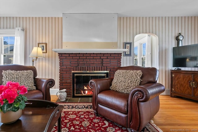 living room with a brick fireplace, wallpapered walls, and wood finished floors