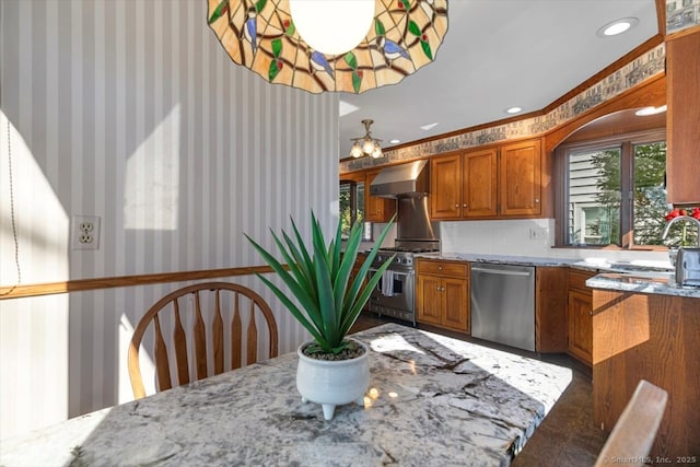 kitchen with light stone countertops, brown cabinetry, wallpapered walls, appliances with stainless steel finishes, and wall chimney exhaust hood