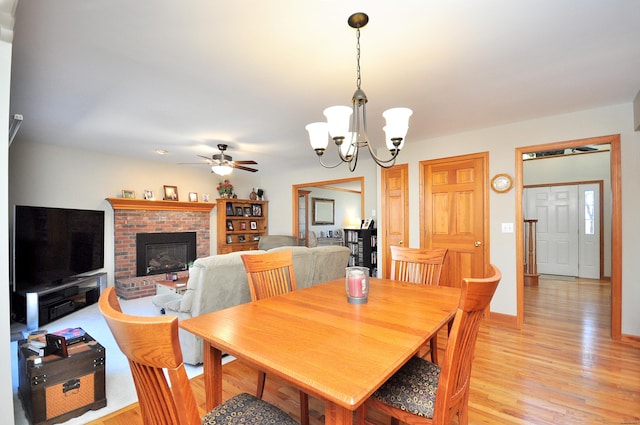 dining space featuring light wood-style flooring, a brick fireplace, ceiling fan with notable chandelier, and baseboards