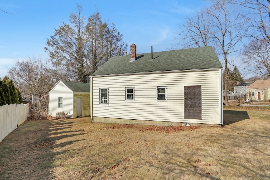 back of house featuring fence, roof with shingles, a chimney, an outdoor structure, and a lawn