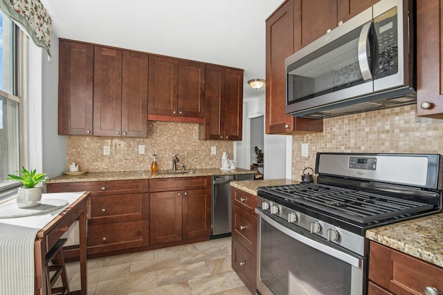 kitchen with stone finish floor, a sink, light stone counters, backsplash, and stainless steel appliances