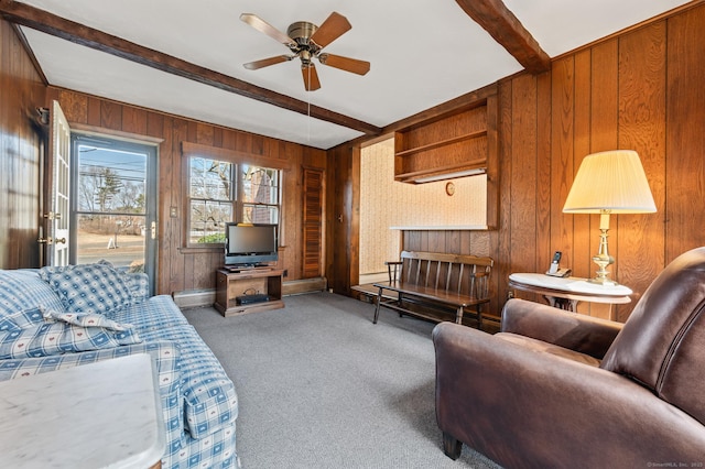 carpeted living room with beamed ceiling, wood walls, and a ceiling fan