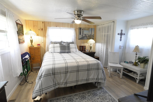 bedroom with wooden walls, a textured ceiling, light wood-style flooring, and a ceiling fan