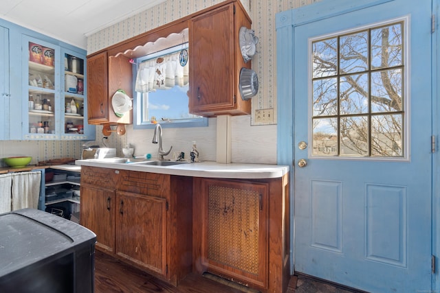 kitchen with brown cabinetry, wallpapered walls, a sink, light countertops, and glass insert cabinets