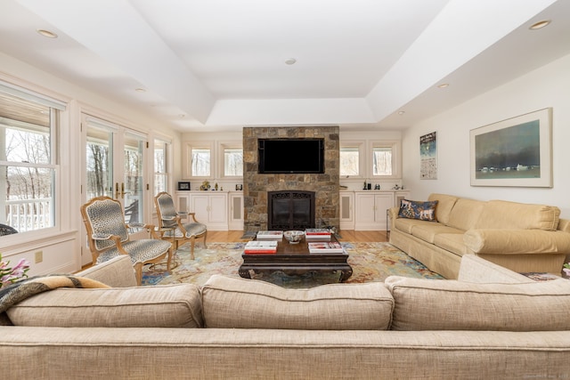 living area with a wealth of natural light, a stone fireplace, a raised ceiling, and wood finished floors