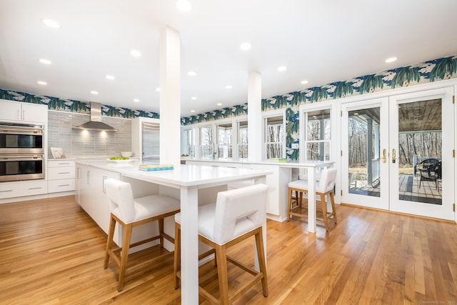 kitchen featuring light wood finished floors, wall chimney range hood, light countertops, french doors, and white cabinetry