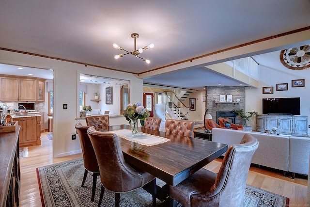 dining room featuring light wood-style floors, a stone fireplace, an inviting chandelier, crown molding, and stairs