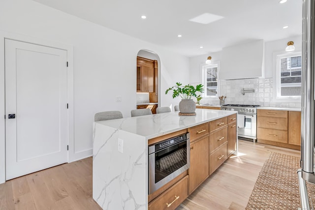 kitchen featuring premium range hood, a center island, arched walkways, stainless steel stove, and light wood finished floors