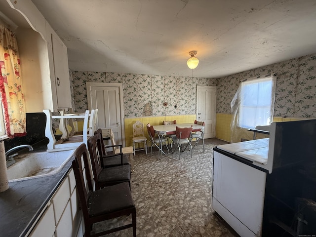 kitchen with wallpapered walls, a sink, tile counters, white cabinets, and wainscoting