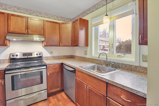 kitchen with light wood finished floors, under cabinet range hood, stainless steel appliances, a textured ceiling, and a sink